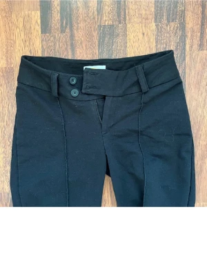 Svarta kostymbyxor från Gina tricot - Snygga svarta kostymbyxor från Gina Tricots  med låg midja och två knappar framtill. De har dekorativa sömmar längs benen och diskret logga på låret. Stretchigt material som sitter skönt och formar sig efter kroppen. Perfekta för dig som vill ha både stil och komfort. Passer mig som har xs/s