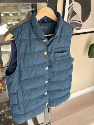 Blå dunväst från Stenströms - Snygg blå dunväst från Stenströms med quiltad design och vita knappar. Västen har ståkrage, bröstficka och två sidofickor. Yttermaterialet har denimlook och insidan är mörkblå. Perfekt för lager-på-lager-stil.