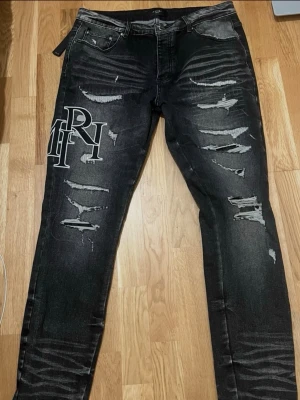 Svarta ripped jeans från Amiri - Säljer ett par svarta jeans från Amiri med stora vita logotryck på vänster lår och flera slitningar och ripped detaljer framtill. Jeansen har en slim fit och är tillverkade i denim med snygg tvättad look. Perfekta för dig som gillar streetwear och statement-plagg.