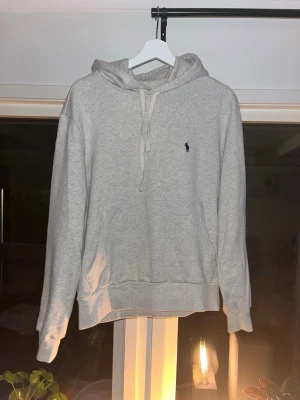 Grå hoodie från Ralph Lauren - Klassisk ljusgrå hoodie från Ralph Lauren med huva och dragsnören. Har en broderad logga på bröstet och en stor magficka framtill. Perfekt för en avslappnad och stilren look.