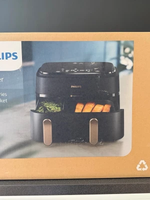 Philips airfryer med två fack - Philips airfryer i svart med två separata fack och digital touchpanel på ovansidan. Utrustad med stilrena handtag i silverfärgad metall och robust plastkonstruktion. Perfekt för att tillaga olika rätter samtidigt med modern teknik.