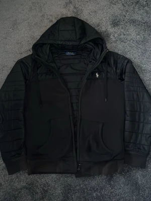 Svart hoodiejacka från Polo Ralph Lauren - Svart hoodiejacka från Polo Ralph Lauren med quiltade detaljer på ärmar och huva. Jackan har dragkedja framtill, två fickor och en broderad logga på bröstet. Tillverkad i polyester och har en sportig, casual look.