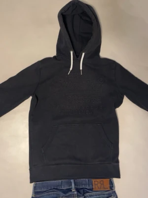 Svart Superdry hoodie - Snygg svart hoodie från Superdry med broderad logga och japansk text på bröstet. Riktigt snygg om du frågar mig. Skriv vid frågor eller funderingar. Väldigt snabba svar 🙂