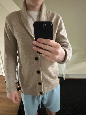 Beige stickad tröja Hollister  - Snygg beige kofta från Hollister med sjalkrage och stora svarta knappar. Koftan har två fickor framtill och ribbade muddar vid ärmslut och nederkant. Perfekt för lager på lager och enkel att matcha med olika outfits.