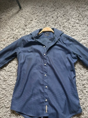 Blå slim fit skjorta från Riley - Snygg blå skjorta från Riley i slim fit-modell. Skjortan har ett diskret rutigt mönster, klassisk krage och vita knappar framtill. Perfekt för dig som gillar en stilren look med lite extra detalj.