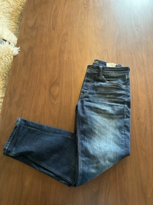 Blå raka jeans från Jack & Jones - Säljer ett par klassiska blå jeans från Nudie Jeans med rak passform och fem fickor. Använda 4 gånger tidigare. Jeansen har en snygg tvätt med lätt slitning framtill och detaljer i kontrastsömmar. Tillverkade i bomull med läderpatch bak i midjan.