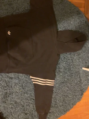 Svart Adidas hoodie med vita ränder - Svart hoodie från Adidas med klassiska vita ränder på ena ärmen och Adidas-logga på bröstet. Stor känguruficka framtill och huva. Perfekt för en avslappnad och sportig stil.