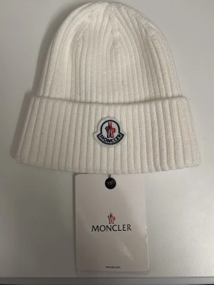 Vit ribbstickad mössa från Moncler - Snygg vit ribbstickad mössa från Moncler med uppvikt kant och klassisk logotyp framtill. Perfekt accessoar för kalla dagar och riktigt trendig stil. Materialet är mjukt och värmande, och mössan har en clean look som passar till det mesta.