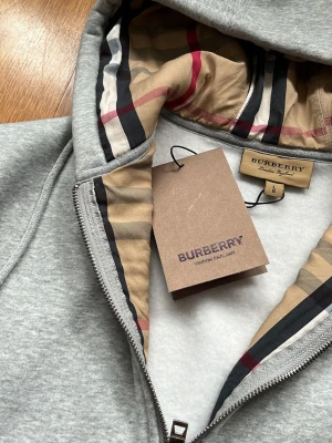 Grå hoodie från Burberry med dragkedja - Snygg grå hoodie från Burberry med dragkedja och klassiskt rutigt foder i huvan. Broderad Burberry-logga på bröstet och exklusiva detaljer. Tillverkad i mjukt material som känns lyxigt och bekvämt. Perfekt för dig som vill ha något stilrent men ändå sticker ut.