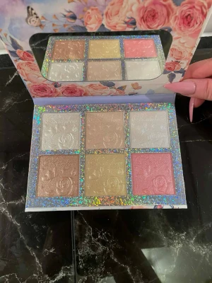 Highlighterpalett med sex nyanser - Säljer en highlighterpalett med sex skimrande nyanser i vitt, guld, beige, rosa och champagne. Medföljer borstar. Paletten har ett holografiskt glitterlock, spegel och vackert blommigt motiv. Perfekt för att skapa glow och highlighta ansiktet.