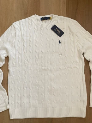 Vit kabelstickad tröja Polo Ralph Lauren - Kabelstickad vit tröja från Polo Ralph Lauren med rund halsringning och diskret broderad logga på bröstet. Tröjan har långa ärmar och ribbade muddar vid ärmslut och nederkant. Perfekt för en clean och klassisk look.