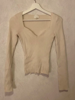 Ljusbeige ribbad topp från H&M - Supersnygg ljusbeige ribbad topp från H&M med lång ärm och v-ringad, fyrkantig urringning. Figurnära passform och stretchigt material som sitter perfekt. Perfekt att styla med jeans eller kjol för en trendig look.