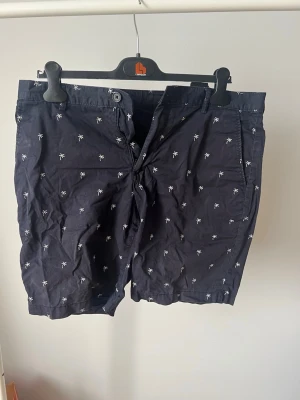 Mörkblå shorts med palmtryck - Snygga mörkblå shorts med små vita palmer tryckta över hela tyget. Klassisk passform med knapp och dragkedja framtill samt fickor både fram och bak. Perfekta för sommardagar och strandhäng.