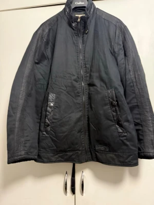 Svart jacka från Laredo med detaljer från DIESEL - Svart jacka från Laredo med ståkrage och dragkedja framtill. Jackan har två snedställda fickor med knappar och quiltade detaljer. Materialet är syntetiskt och jackan har diskreta mönster längs ärmar och rygg.