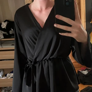 Svart omlottblus från Chica London - Snygg svart omlottblus från Chica London med långa ärmar och knyt i midjan. Blusen har v-ringning och ett flowy tyg som ger en elegant look. Perfekt för dig som gillar stilrena och klassiska plagg.