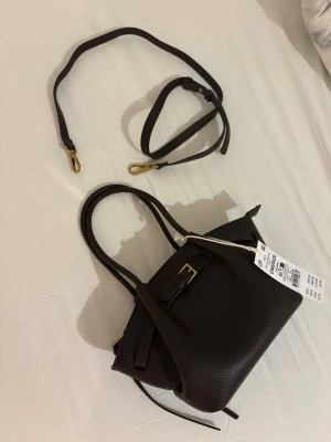 Brun minihandväska med avtagbar rem - Snygg brun minihandväska från GinaTricot i strukturerat skinnimitation. Väskan har två handtag, avtagbar axelrem och dekorativt spänne framtill i guld. Perfekt för att bära det viktigaste och ge din outfit en trendig touch.