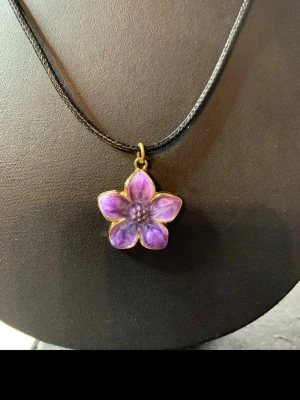 Lila blomformad halsbandshänge - Snyggt halsband med ett lila blomformat hänge i guldtonad ram. Hänget har fem kronblad och små pärldetaljer i mitten. Hängs på ett svart snöre. Perfekt accessoar för att lyfta din outfit med en färgklick och unik design.