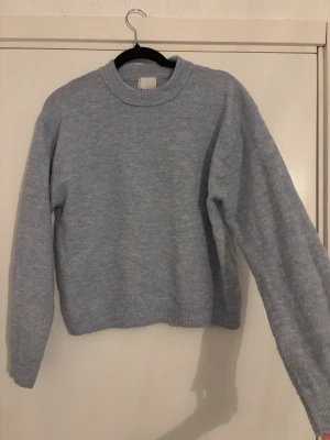 Ljusgrå stickad tröja med rund hals - En stilren ljusgrå stickad tröja med rund halsringning och långa ärmar. Tröjan har ribbstickade muddar vid ärmslut och nederkant, vilket ger en klassisk och enkel look. Perfekt att matcha med jeans eller kjol för en avslappnad stil.