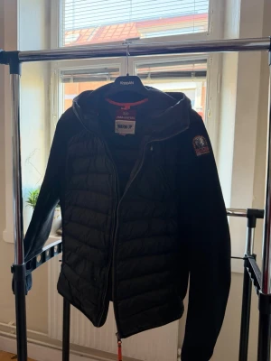 Parajumper cardigan - Snygg svart pufferjacka från Parajumler med quiltad framsida och rygg, samt mjuka ärmar. Jackan har huva, dragkedja framtill och logotyp på ärmen. Perfekt för kalla vinterdagar och riktigt skön att bära.