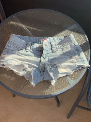 Ljusblå jeansshorts med fransar - Säljer ett par ljusblå jeansshorts med råa, fransiga kanter och dekorativa nitar på bakfickorna. Klassisk femficksmodell med knapp och bälteshällor. Perfekta för varma dagar och en avslappnad stil.⭐️
