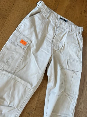 Beiga cargobyxor från Obey  - Snygga beiga cargobyxor från Obey med flera praktiska fickor på sidorna och en orange logodetalj. Byxorna har raka ben och är tillverkade i ett slitstarkt bomullsmaterial. Perfekta för en avslappnad och trendig look.