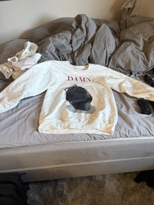 Vit DAMN. sweatshirt med tryck - Vit sweatshirt med röd text 'DAMN.' framtill och ett stort svart tryck av ett huvud. På baksidan står det 'Love's Gonna Get You Killed' i svart text. Tröjan har rund halsringning och långa ärmar. Perfekt för dig som gillar streetwear och statement-plagg.