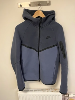 Storleken är XS men den sitter som S/Nike Tech Fleece hoodie blågrå - Nike Tech Fleece hoodie i blågrå nyans med svart Nike-logga på bröstet. Dragkedja framtill, huva och fickor på sidorna samt en extra ficka med dragkedja på ärmen. Sportig och stilren design i mjukt fleecematerial.