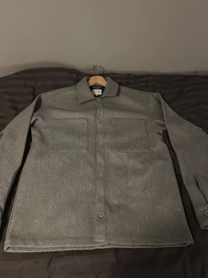 Grå overshirt från Jack & Jones - Stilren grå overshirt från Jack & Jones i storlek XS. Skjortjackan har klassisk krage, raka ärmar, knäppning framtill och två stora bröstfickor. Materialet är mjukt och ger en clean look som passar till många outfits.