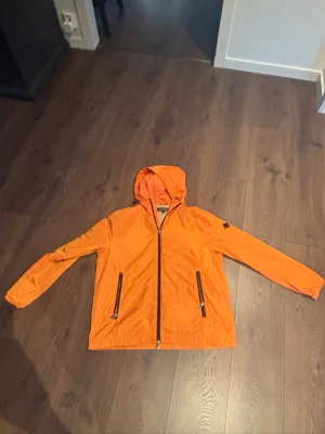 J.linberg jacka  - Snygg orange vindjacka med huva och svarta dragkedjor framtill samt på fickorna. Jackan är lätt och har en sportig look, perfekt för blåsiga dagar. Justerbar huva och elastiska ärmslut ger en bekväm passform.