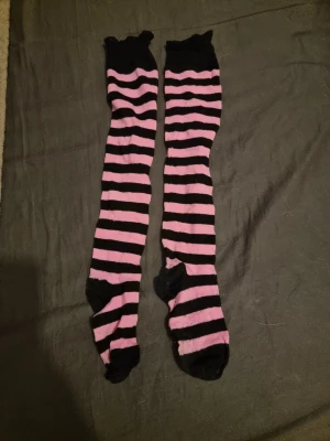 Randiga knästrumpor i rosa och svart - Säljer ett par långa knästrumpor med breda rosa och svarta ränder. Strumporna har svart tå och häl, och är perfekta för dig som vill sticka ut med en färgglad och lekfull stil. Passar till både kjol och shorts.