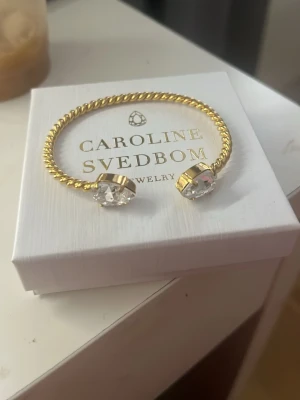 Guldarmband med kristaller Caroline Svedbom - Snyggt armband i guldton från Caroline Svedbom med tvinnad design och två stora, klara kristaller i ändarna. Perfekt accessoar för att lyfta din outfit med lite extra bling och elegans. Aldrig använd eftersom jag redan har en i silver, pris går att diskutera❤️