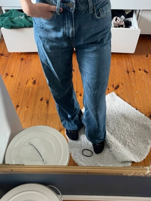 Blå raka jeans med hög midja - Säljer ett par klassiska blå jeans med rak passform och hög midja. Jeansen har fem fickor och stängs med dragkedja och knapp. Tillverkade i ett kraftigt denimtyg som ger en tidlös look. Perfekta till vardags och kan stylas på många sätt.