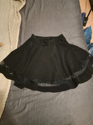 Svart kortkjol med meshdetalj - Säljer en svart kortkjol med volang och meshdetalj längs kanten. Kjolen har en utställd form och bred midjeresår för skön passform. Perfekt för dig som vill ha en enkel men snygg kjol till garderoben. Vet inte vilken storlek men skulle gissa på M.