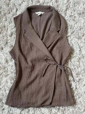 Brun omlottväst från H&M - Säljer en brun omlottväst från H&M i storlek XS. Västen har en krage, knytband i midjan och är gjord i ett strukturerat, våfflat tyg av 100% polyester. Perfekt för att addera lager till din outfit och ge en chill men ändå stilsäker vibe.