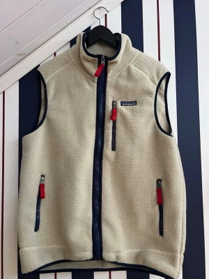 Patagonia väst - Beige fleeceväst från Patagonia med marinblå detaljer och röda dragkedjor.  Mjuk och väldigt skön. Har ägt den i nån månad men kommer inte till användning mycket alls så säljer den. Finns inga tydliga spår av användning.