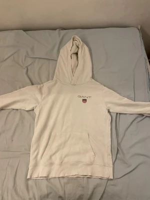 Vit hoodie från GANT med logga - Snygg vit hoodie från GANT med broderad logga och sköld på bröstet. Klassisk känguruficka framtill och ribbade muddar. Perfekt för en chill och stilren look. Tillverkad i mjukt material för extra komfort. Är lite trasig om ärmarna 