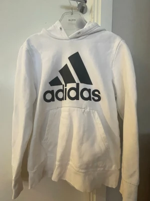 Vit hoodie från Adidas med logga - Snygg vit hoodie från Adidas med stor svart logga på bröstet. Klassisk känguruficka framtill och huva med dragsko. Mjuk insida i fleece och ribbade muddar vid ärmar och nederkant. Perfekt för en sportig och avslappnad stil.
