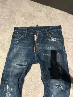 DSQUARED2 Slim jeans med dragkedja - Säljer ett par äkta blå DSQUARED2 jeans i skinny fit med coola slitningar, färgstänk och en stor dragkedja framtill. Jeansen har klassisk femficksdesign och är gjorda i bomull. Perfekt för dig som gillar streetstyle och vill sticka ut. 
