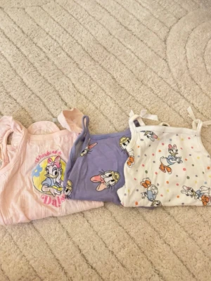 Söta toppar med Disney-tryck - Tre olika toppar med motiv av Kajsa Anka i pastellfärger. En rosa t-shirt, ett lila linne och ett vitt linne med färgglada prickar och tryck. Alla har gulliga detaljer. De är linnebodys.