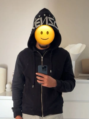 Svart Kenzo hoodie med dragkedja - Svart hoodie från Kenzo med vit logga 'KENZO PARIS' tryckt på huvan. Tröjan har dragkedja framtill, dragsko i huvan och ribbade muddar vid ärmslut och nederkant. Perfekt för en stilren och trendig look. 