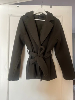 Olivgrön kappa från Glamorous - Stilren olivgrön kappa från Glamorous med knytskärp i midjan och breda slag. Kappan har en rak passform och är fodrad på insidan. Perfekt för dig som vill ha en klassisk look med modern touch.