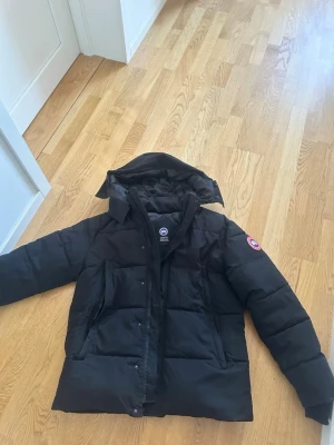 Svart dunjacka från Canada Goose - Säljer en svart dunjacka från Canada Goose med avtagbar huva och klassisk logotyp på ärmen. Jackan har dragkedja och knappar framtill samt två stora fickor. Perfekt för kalla vinterdagar och riktigt varm tack vare dunfyllningen. Drag kjedjan fungerar inte men det går att stänga med knapparna utan problem 👍 