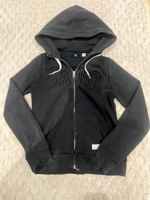 Svart hoodie med dragkedja från GANT - Säljer en svart hoodie från GANT i storlek XS. Hoodien har dragkedja framtill, stor huva och fickor på magen. GANT-loggan är broderad i svart på bröstet. Tillverkad i mjukt bomullsmaterial, perfekt för en avslappnad stil.