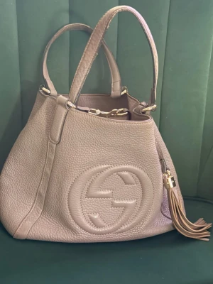 Grå Gucci Soho handväska i läder - Säljer en Grå Gucci Soho handväska i mjukt, kornigt läder med stora GG-loggan framtill. Väskan har två handtag, guldfärgade metalldetaljer och en dekorativ tofs. Perfekt för dig som gillar lyxiga accessoarer med klassisk design.