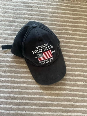 Svart Vinson Polo Club keps - Snygg svart keps från Vinson Polo Club med broderad amerikansk flagga och vit text framtill. Justerbart spänne bak för perfekt passform. Klassisk böjd skärm och ventilerande hål. Tillverkad i bomullstyg som känns bekvämt och stilrent.