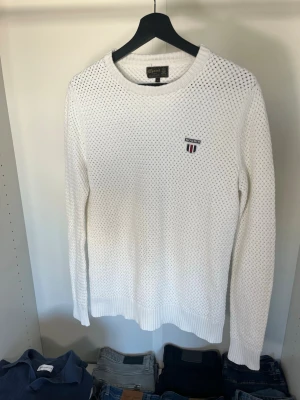 Vit stickad tröja från Morris - Vit stickad tröja från Morris med rund halsringning och en logotyp på bröstet. Tröjan har ett snyggt hålmönster och ribbade muddar vid ärmslut och nederkant. Perfekt för en stilren och klassisk look.