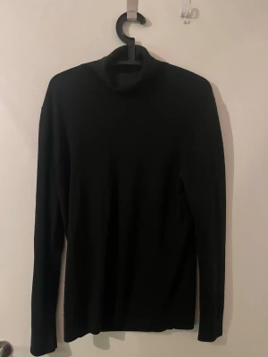 Svart polotröja i stickat material - Säljer en svart stickad polotröja med lång ärm och klassisk passform. Tröjan har en hög krage och är perfekt för lager-på-lager-stil. Enkel och stilren design som funkar till det mesta.