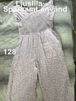 Ljuslila blommig jumpsuit - Supersöt ljuslila jumpsuit med vita små blommor och smock upptill. Vida ben och korta ärmar ger en avslappnad och trendig look. Perfekt för dig som gillar pasteller och blommigt mönster.🌸 sparsamt använd . Storlek 128