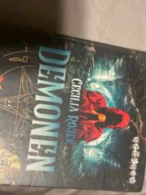 Demonen av Cecilia Rojek - En spännande skönlitterär bok med titeln 'Demonen' av Cecilia Rojek. Omslaget är mörkt med en mystisk figur i röd kåpa och magiska symboler, vilket ger boken en fantasy- och äventyrskänsla. Perfekt för dig som gillar övernaturliga berättelser.