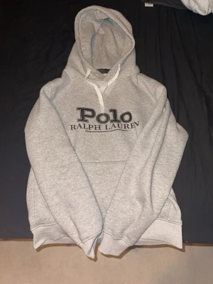 Grå hoodie från Polo Ralph Lauren - Snygg grå hoodie från Polo Ralph Lauren med stor logga broderad på bröstet. Klassisk känguruficka framtill och vit dragsko i huvan. Tillverkad i mjukt bomullsmaterial som känns skönt mot huden. Perfekt för en avslappnad och stilren look.